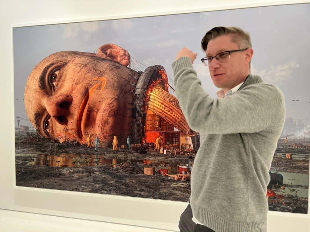A digital NFT artwork displayed in a gallery showing a colossal human head resembling a tech billionaire lying in a dystopian industrial landscape, with a contemporary digital artist standing beside the artwork and pointing at it.
